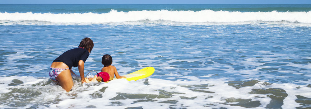 Kleiner Junge auf dem Surfbrett liegend mit seinem Au-pair im Wasser